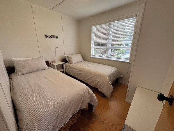 A bedroom with two beds, a small bedside table with a lamp, and a window with blinds.