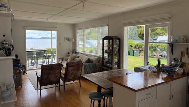 A spacious kitchen with a wooden countertop, a dining area with chairs, a sofa, and a view of a lake through large windows. - Lake Rotoiti Lakefront Bach with Stunning Sunsets (Rotoiti Forest)