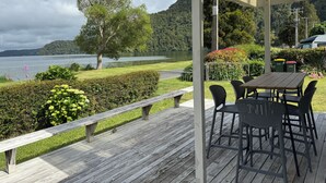 An outdoor deck with a table and chairs, overlooking a lake and forested hills. - Lake Rotoiti Lakefront Bach with Stunning Sunsets (Rotoiti Forest)