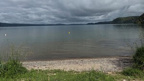 A calm lake with a pebbled shore and overcast skies. - Lake Rotoiti Lakefront Bach with Stunning Sunsets (Rotoiti Forest)