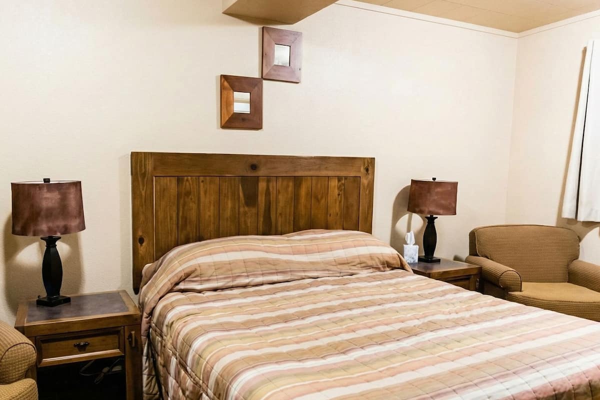Camera d'albergo con una testiera in legno, un letto grande con biancheria a righe, due comodini con lampade, una sedia e un piccolo specchio a parete.