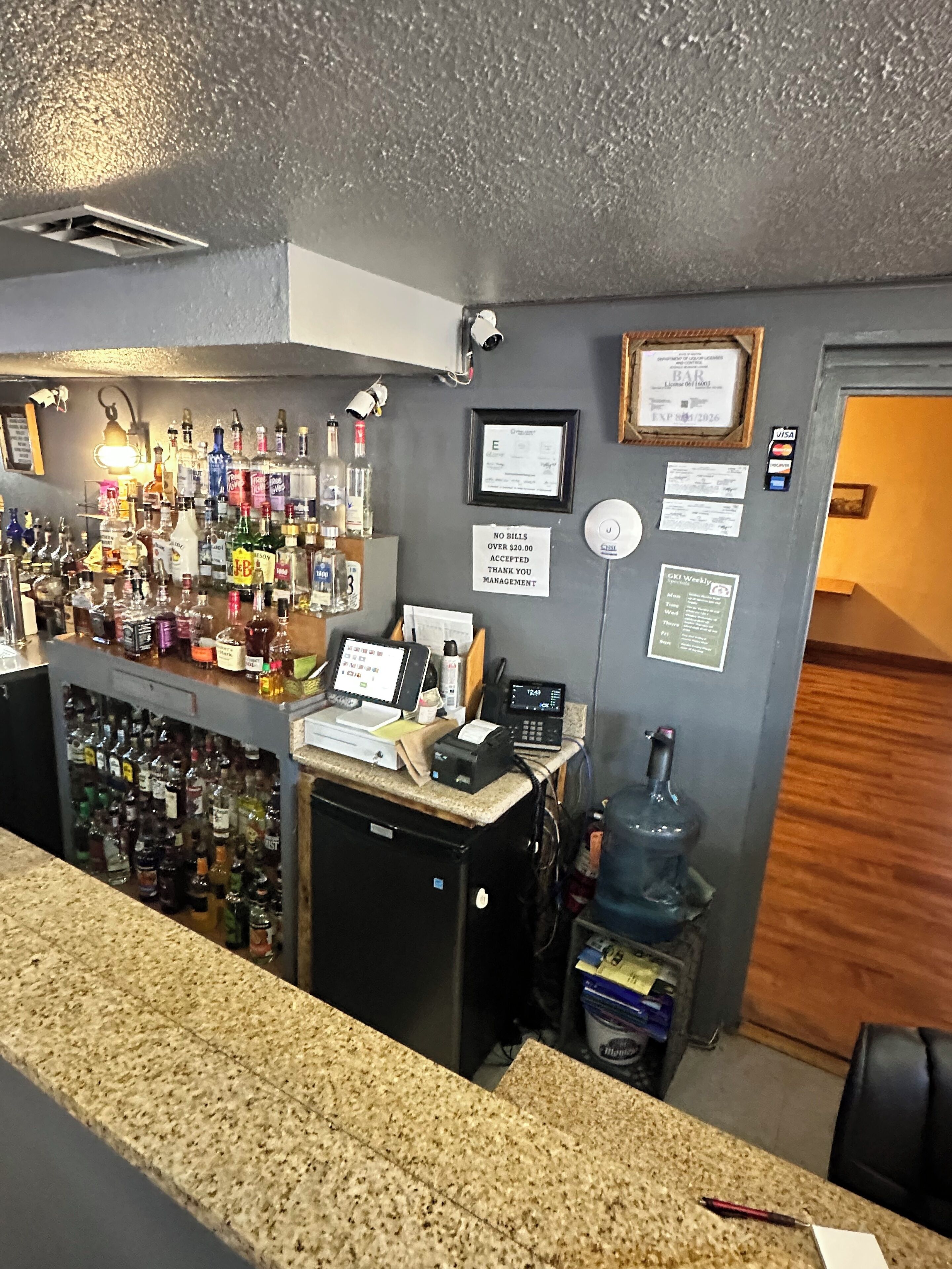 Zona de bar con mostrador, varias botellas de licor, una caja registradora y un reloj digital.