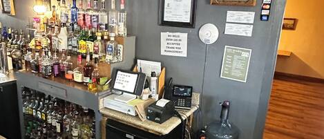 Zona de bar con mostrador, varias botellas de licor, una caja registradora y un reloj digital.