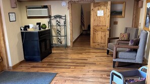 A hallway with wooden flooring, a blue doormat, a metal cart with bags, a wooden door, and a framed picture on the wall. - General Kearny Inn (Kearny)