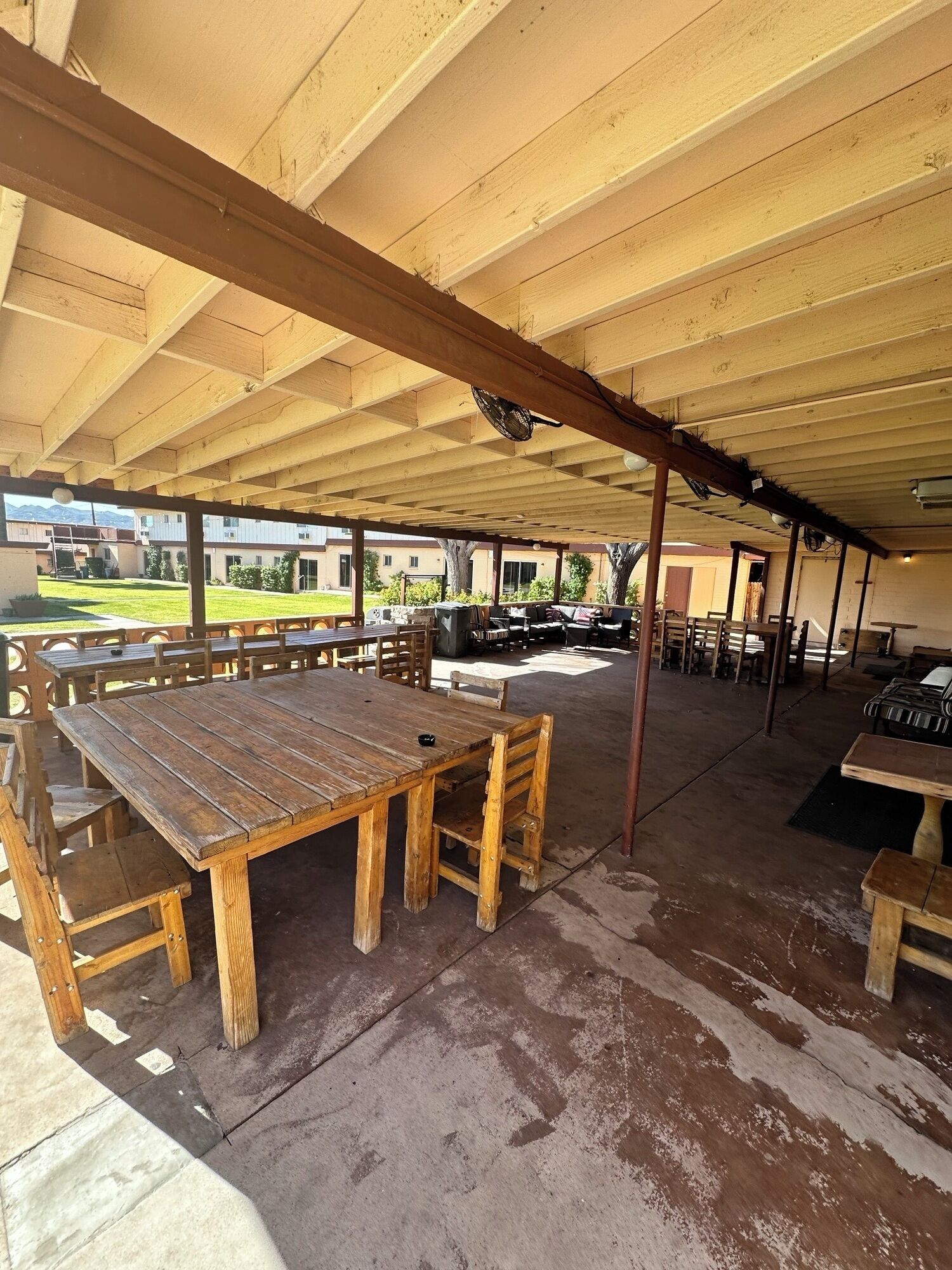 Un patio techado al aire libre con mesas y bancos de madera, un ventilador y vista a una zona residencial al fondo.
