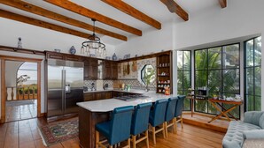 Une cuisine moderne avec des poutres en bois, un grand îlot central doté de tabourets de bar et une vue sur la verdure grâce à de grandes fenêtres.