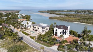 Une grande maison blanche à deux étages, dotée d’une piscine, entourée d’une végétation tropicale et bordée par une rivière.
