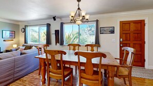 A dining area with a wooden table and chairs, a chandelier, and a view through large windows. - Charming Fort Bragg Home with Ping-Pong & Fire Pit (Fort Bragg)