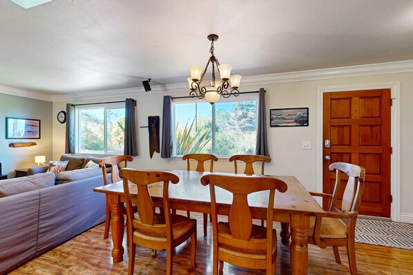 A dining area with a wooden table and chairs, a chandelier, and a view through large windows. - Charming Fort Bragg Home with Ping-Pong & Fire Pit (Fort Bragg)