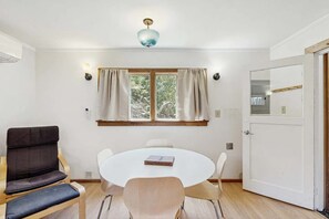 Ein Zimmer mit rundem Tisch, zwei Stühlen, einer Couch, einem Fenster mit Vorhängen, einer Deckenleuchte und einer Tür.