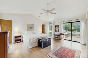 A spacious bedroom with a large bed, a ceiling fan, a wooden dresser, a chest of drawers, a bench, and a large window offering a view of greenery. - Beachfront Bliss with Ocean Views, Tub, & BBQ (Fort Bragg)