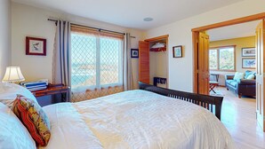 Ein Schlafzimmer mit einem großen Bett, einem Nachttisch mit Lampe, einem Fenster mit Vorhängen, einem Kleiderschrank und Blick ins Wohnzimmer.