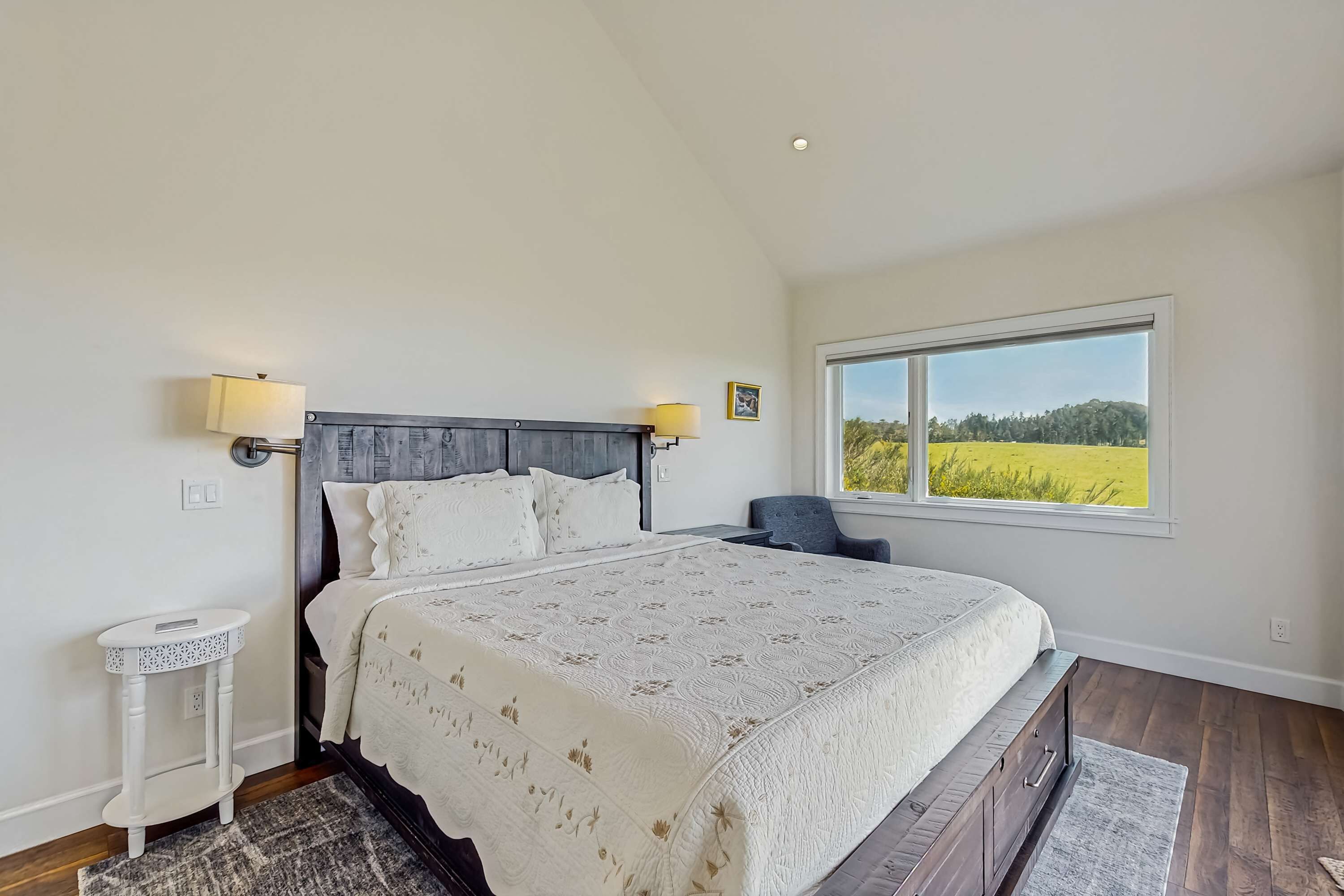 Quarto com uma cama grande, uma mesinha de cabeceira, uma cadeira e uma janela com vista para um campo.