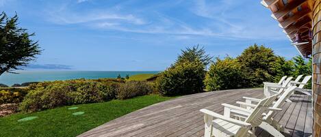 A wooden deck with white Adirondack chairs overlooking a scenic ocean view.