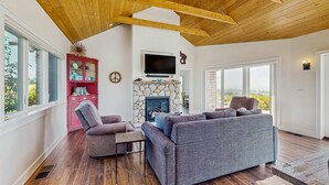 A spacious living room with a stone fireplace, wooden ceiling beams, and large windows offering a view of the outdoors. - Albion Ridge Overlook - Luxury & Modern (Albion)