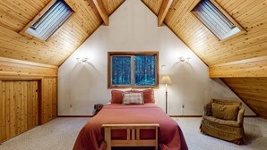 Une chambre avec un plafond en pente, des murs en bois, une grande fenêtre, un lit, une chaise et une lampe.