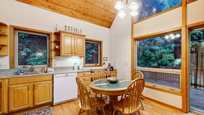 Une cuisine avec des meubles en bois, un lave-vaisselle et un coin repas comprenant une table et des chaises. De grandes fenêtres offrent une vue sur les arbres.