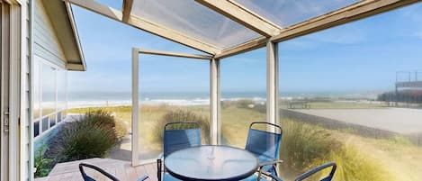 A covered patio with a table and chairs, overlooking a beach and ocean.