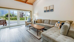 A spacious living room with a large sofa, a coffee table, and a chair. There is a large window with a view of greenery outside.