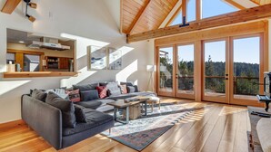 Ein modernes Wohnzimmer mit einem großen grauen Sofa, Holzboden und Blick auf einen Wald durch große Glasfenster.