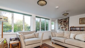 A spacious living room with two beige sofas, a wooden coffee table, and a patterned rug. Large windows provide natural light, and a bookshelf is visible in the background. - Ve-Marn Cottage | Kilrenny (Kilrenny)