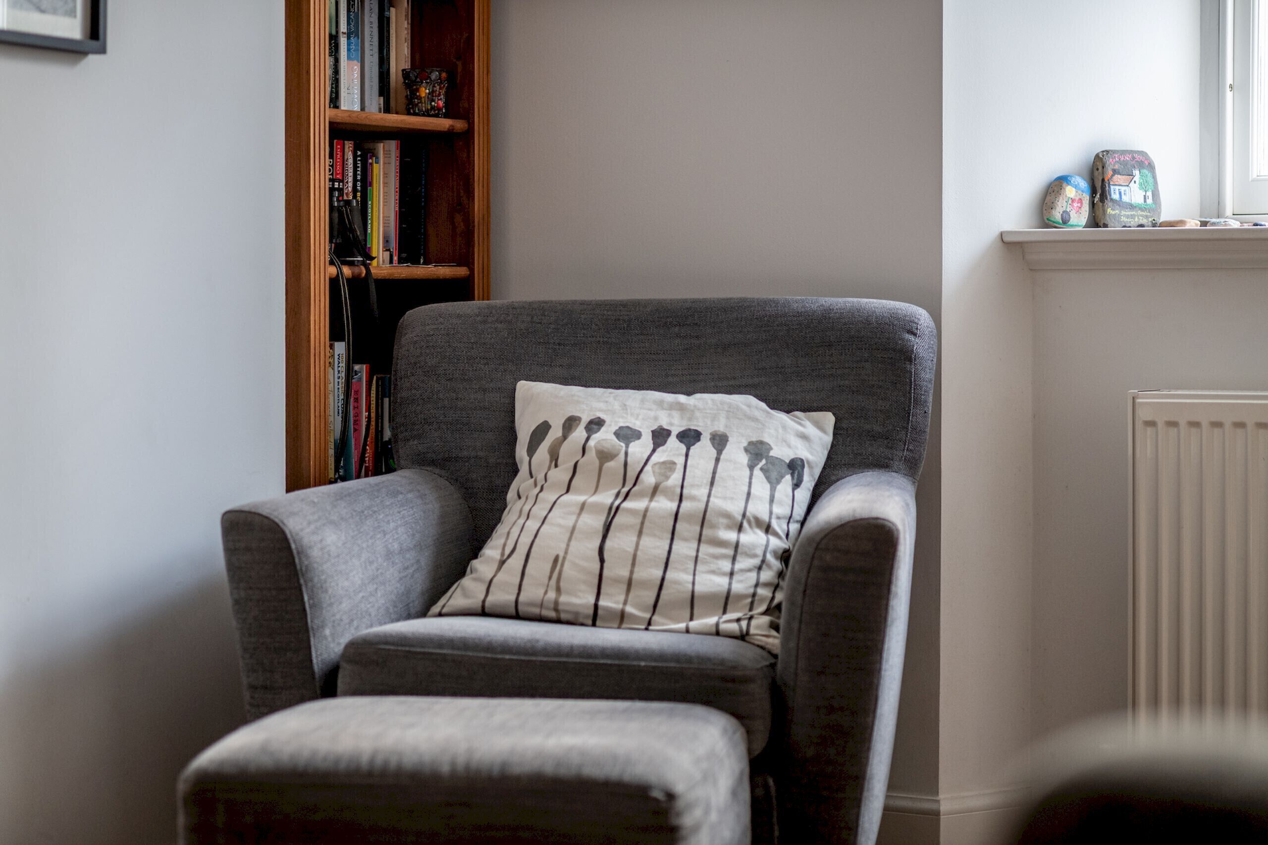 A grey armchair with a white pillow featuring a floral pattern, positioned in a room with a bookshelf and a radiator.