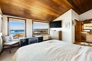 A bedroom with a large bed, wooden ceiling, a desk with a computer, a TV, and a view of the ocean. - Oceanfront Sanctuary - Private Cove, Deck, Grill (Fort Bragg)