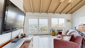 Salón con televisor de pantalla plana, muebles de madera, sofá rosa y vista al océano a través de amplias ventanas.