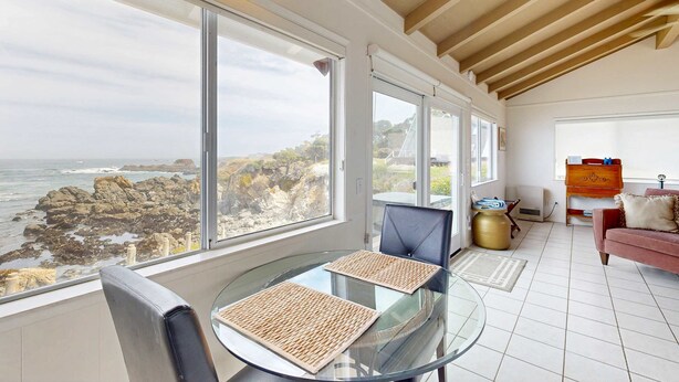 Un salón moderno con ventanas amplias que dan a una costa rocosa, una mesa de vidrio con dos sillas, un sofá rosa y un armario de madera.