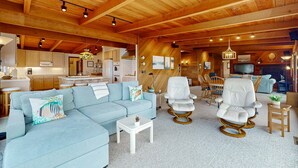 A spacious living area with a light blue sofa, white armchairs, and a small coffee table. A kitchen is visible in the background with wooden cabinets and a dining area with wicker chairs. - Oceanfront Haven with Beach Access, Views & Deck (Fort Bragg)
