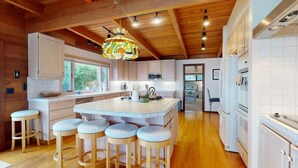 A spacious kitchen with wooden beams, a central island with stools, white cabinets, and a colorful pendant light. - Oceanfront Haven with Beach Access, Views & Deck (Fort Bragg)