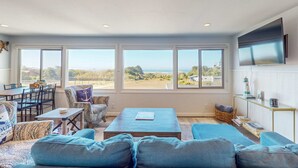 A spacious living room with a large window, a flat-screen TV, and a cozy seating area. - Ocean View Apartment - Beach Access, Yard, Bikes (Fort Bragg)