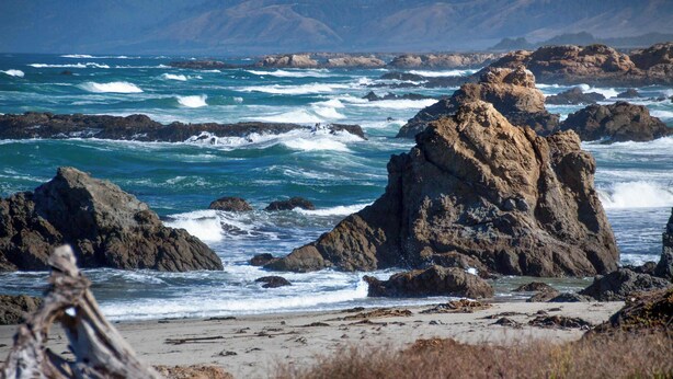 A rocky coastline with waves crashing against large boulders and a sandy beach. - Ocean View Apartment - Beach Access, Yard, Bikes (Fort Bragg)