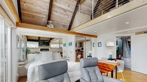 Un interior moderno con una cama, dos sillones grises, una mesa de comedor de madera y una cocina al fondo.