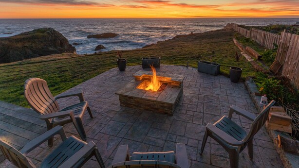 Un espacio al aire libre con fogata, sillas y vista al océano al atardecer.