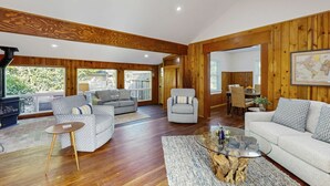 Ein geräumiges Wohnzimmer mit Holzwänden und Holzdielen, einem Kamin, einem Essbereich und großen Fenstern.