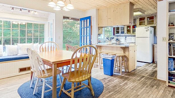 Un coin repas avec une table et des chaises en bois, un tapis bleu et une vue dégagée sur l’extérieur grâce à une grande fenêtre.
