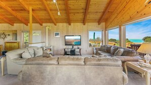 Amplia sala de estar con techo de madera, ventanas grandes con vistas al océano y un televisor de pantalla plana montado en la pared.