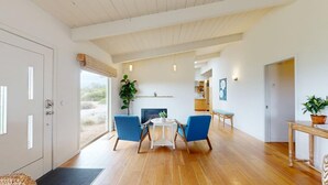 Ein geräumiges Wohnzimmer mit Holzboden, einem zentralen Tisch mit Stühlen, einem Kamin und einem großen Fenster mit Blick ins Freie.