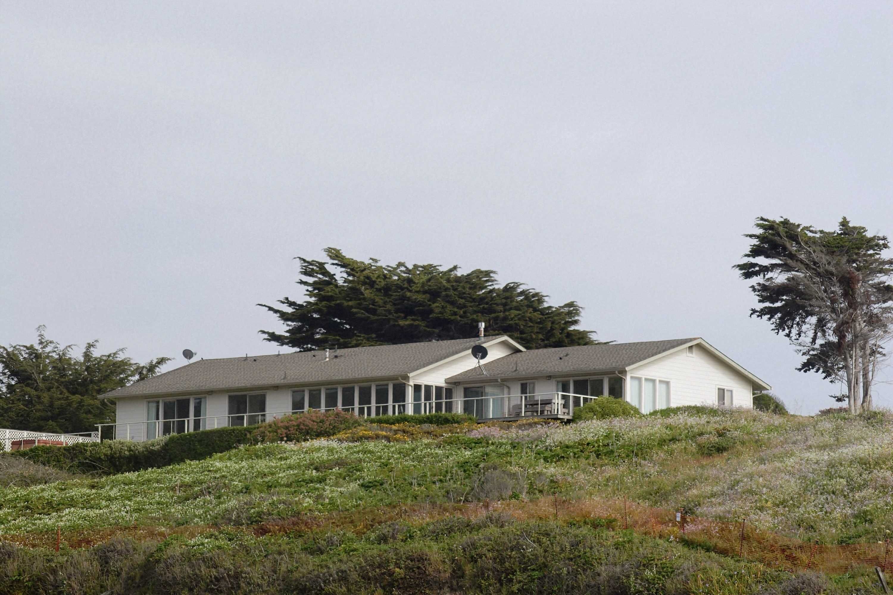 A white house with a long porch, surrounded by a natural landscape with wildflowers and trees.