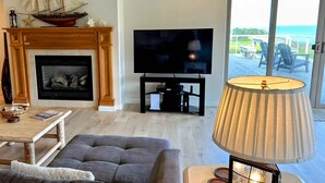 A living room with a fireplace, a television, a lamp, and a view of the ocean through a sliding door. - Oceanfront Haven - Hot Tub & Private Beach Access (Fort Bragg)