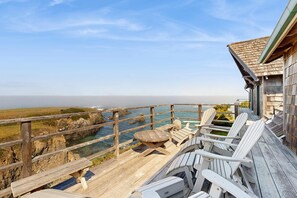 A wooden deck with Adirondack chairs and a wooden table overlooking a coastal landscape with rocky cliffs and a clear sky. - Oceanfront Home - Stunning Views & Private Hot Tub (Fort Bragg)
