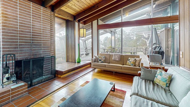 A spacious living room with a fireplace, wooden floors, a sofa, and large windows offering views of the outdoors. - Oceanfront Blufftop Home - Wide Deck & Water Views (Elk)