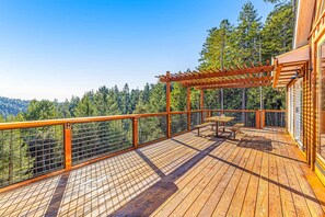 A wooden deck with a pergola, a picnic table, and a railing overlooking a forested area. - Stunning 3BR with ridge top views and hot tub (Little River)