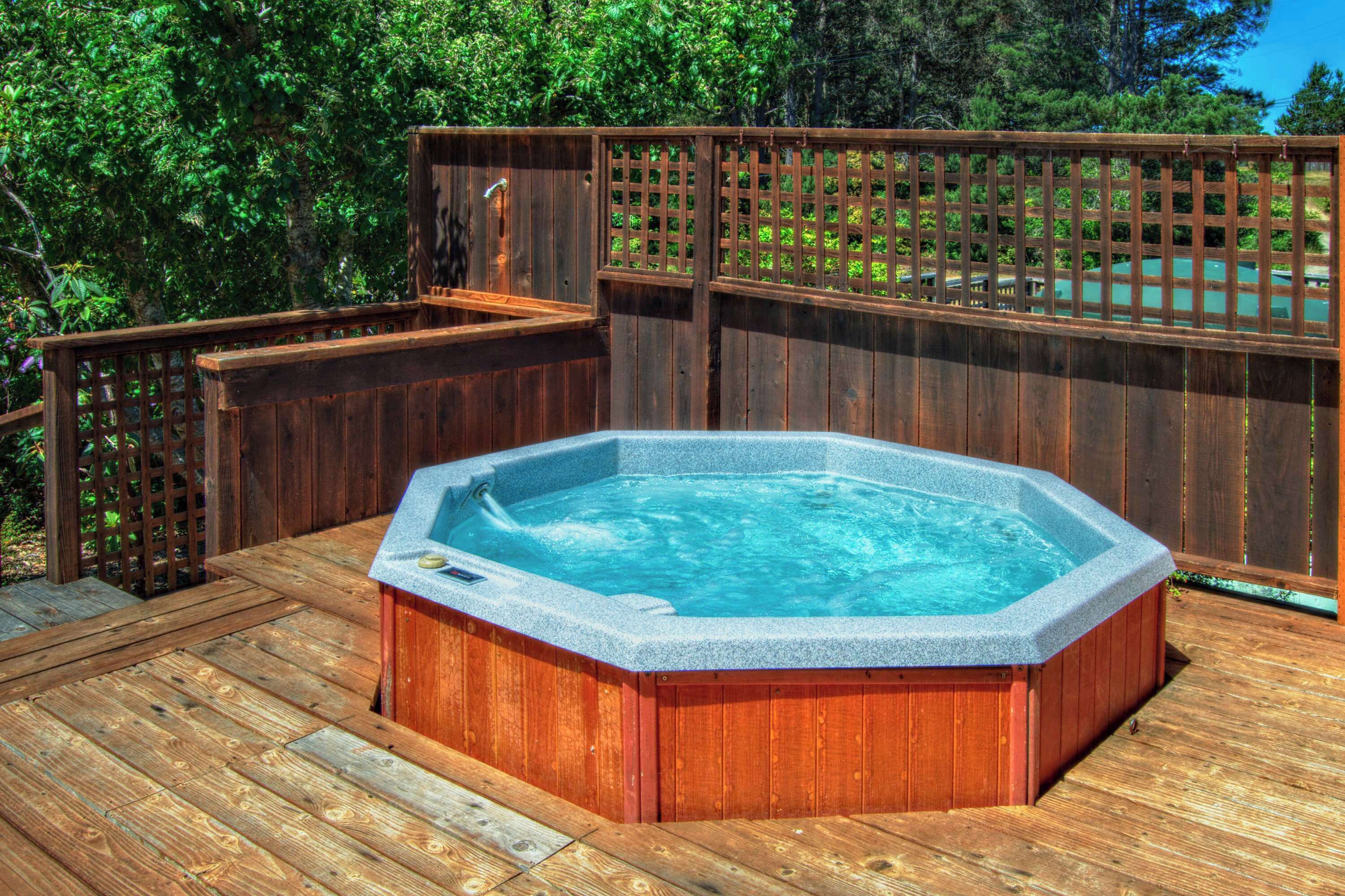 Un jacuzzi en una terraza de madera con valla y follaje al fondo.