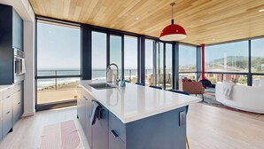 Una cocina moderna con una isla grande, vistas a la playa y un comedor con sofá blanco y lámpara colgante roja.