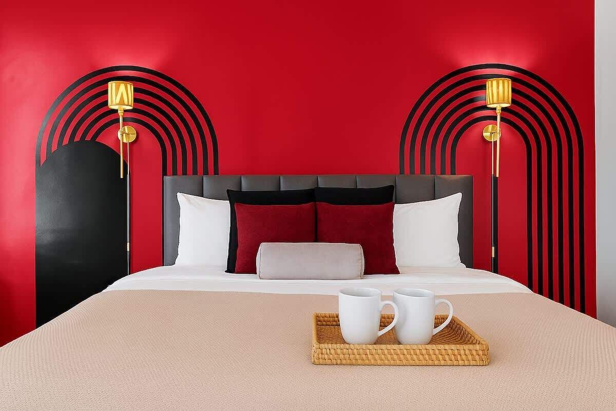 Habitación de hotel con pared roja, cabecero negro y gris, y dos lámparas de noche.