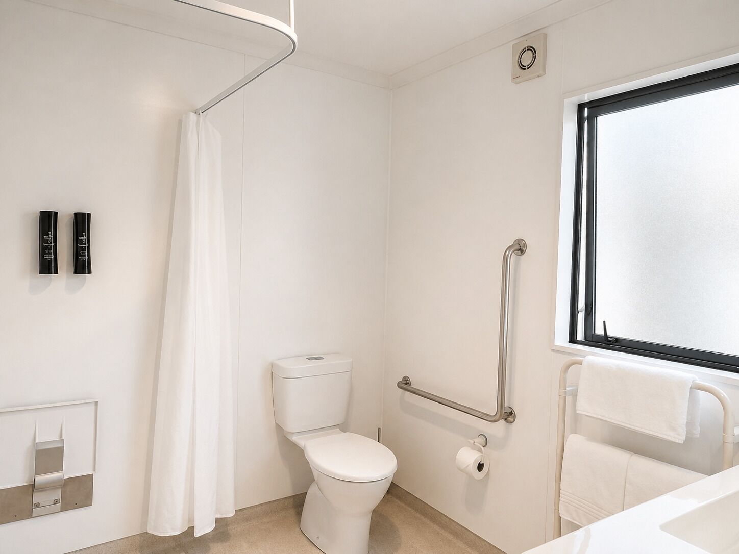 A bathroom with a toilet, a shower curtain, a handrail, a window, and a towel rack.
