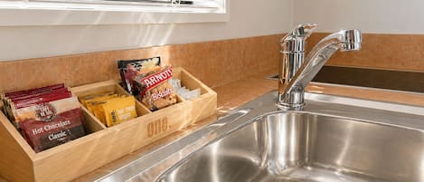 A kitchen with a stainless steel sink, a wooden spice rack with snacks, and a window with blinds.