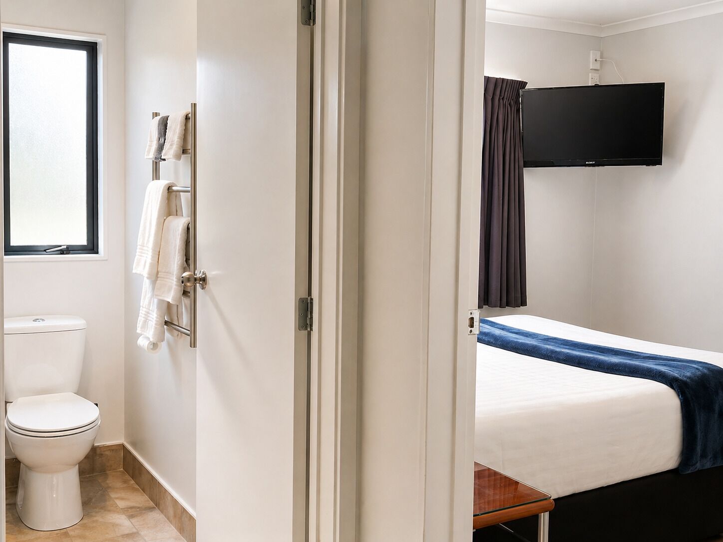 Quarto de hotel com sanita, cama com manta azul, televisão presa à parede e janela com cortinas.